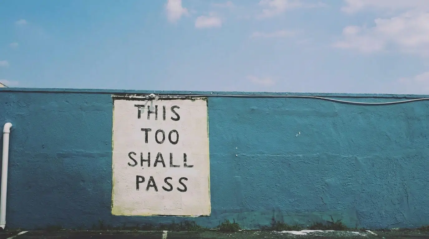 Blue exterior wall with a painted sign reading “This Too Shall Pass.”